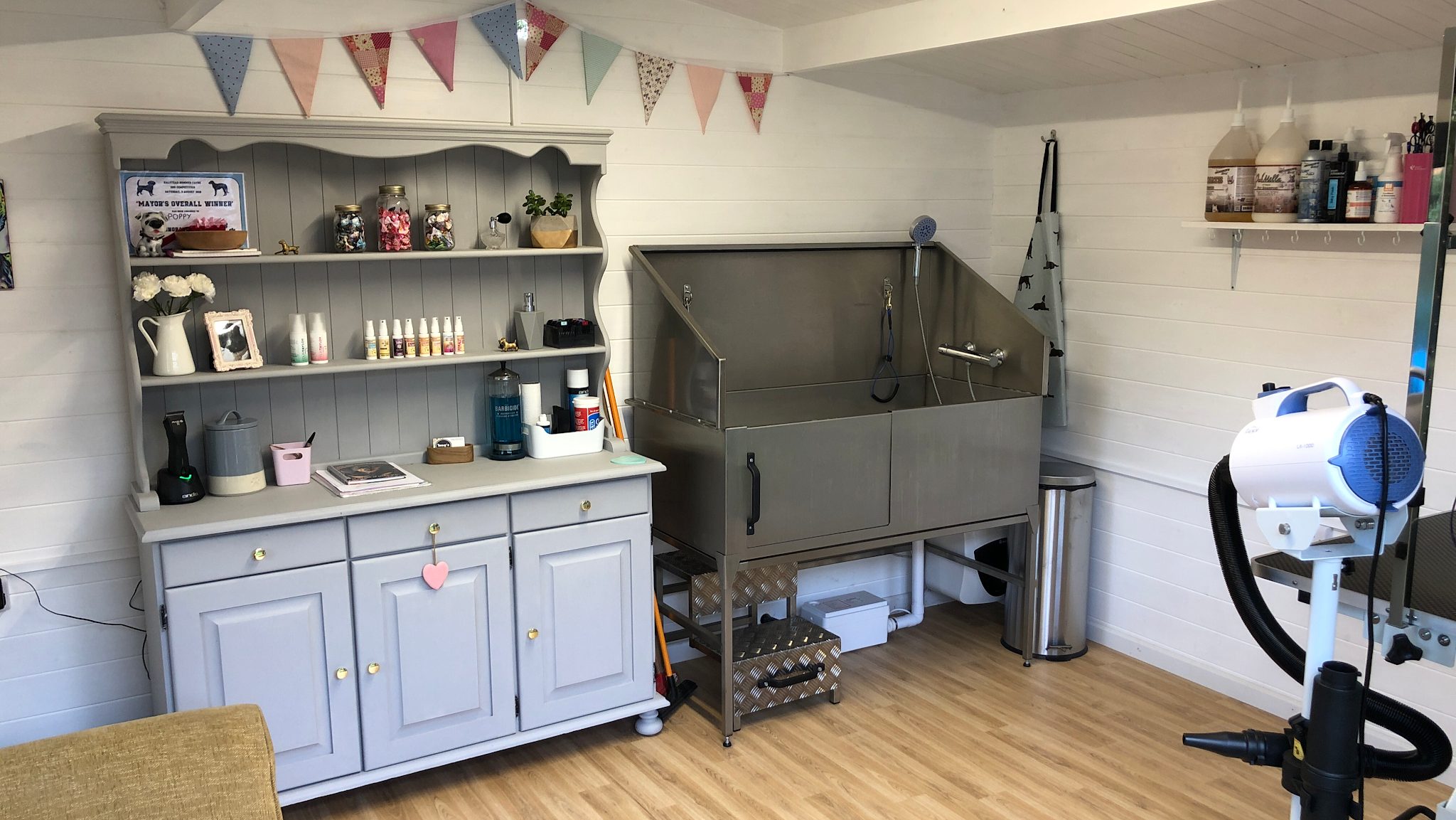 Inside the grooming studio: a grey dresser with jars of bows, bunting, a stainless steel dog bath and a dryer
